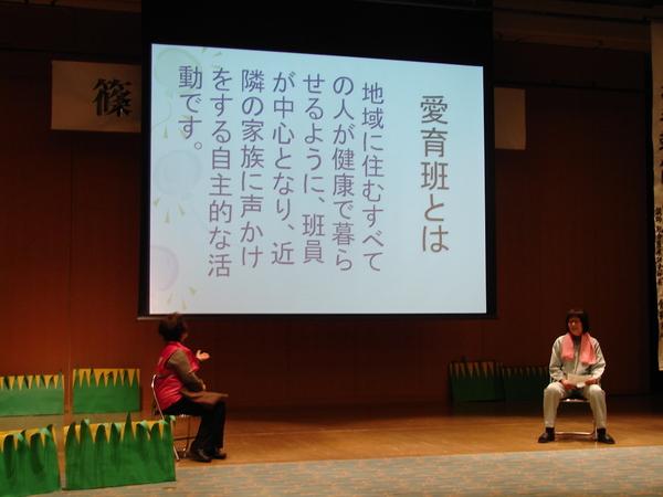 スクリーンに出ている言葉を舞台の上で椅子に座りながら女性が説明している写真